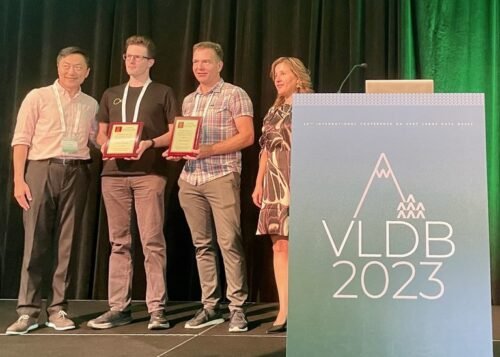 Four people stand on a stage at a conference, with two men proudly holding plaques for the best paper award. A podium on the right displays VLDB 2023. A black curtain serves as the backdrop.