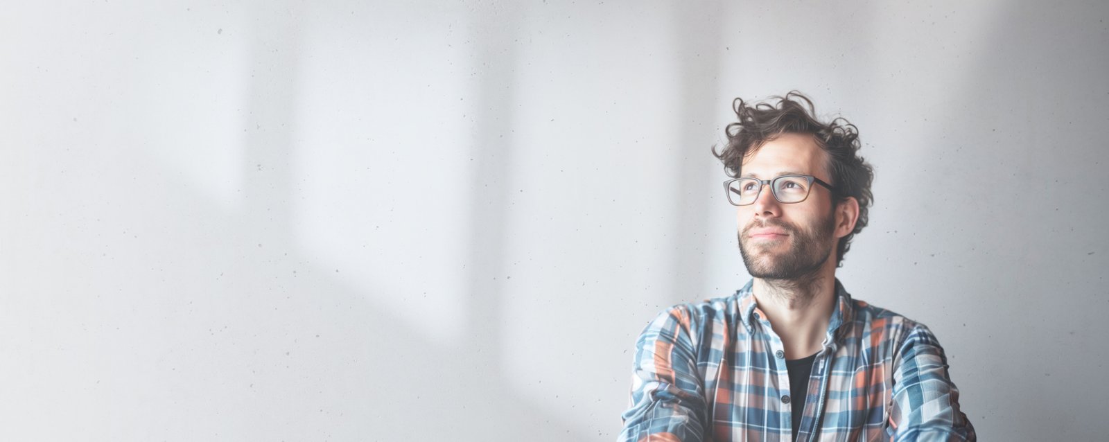 A man with glasses and curly hair is looking thoughtfully to the side. He is wearing a plaid shirt and stands against a plain light gray wall with soft shadows.