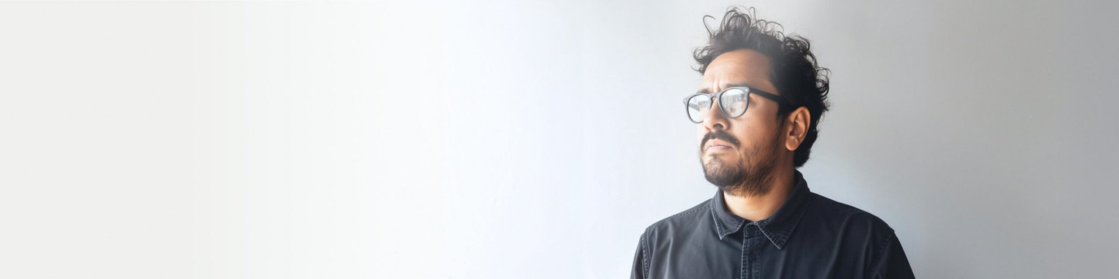 Man wearing glasses and a dark shirt looking contemplatively to the side with a neutral expression against a simple, light-colored background.